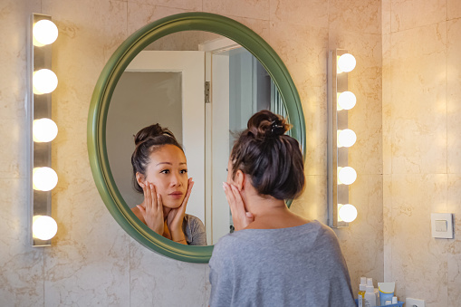 Rear view shot of a worried East Asian woman putting her hands on her cheek, checking her face skin from aging sign. She's standing in front a mirror with lights on both side.