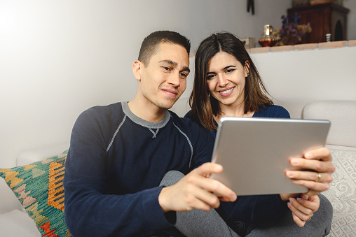 appy young couple at home with electronic device having a video call to friends or family. Smiling and talking sitting on the sofa.