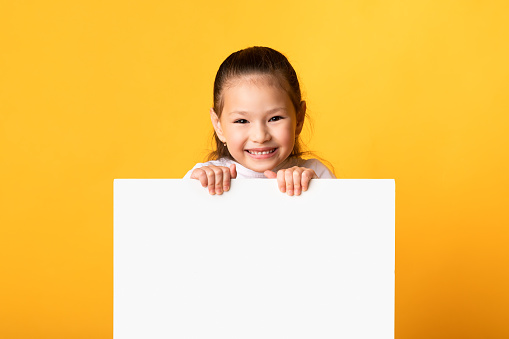 Advertisement Concept. Smiling little girl holding and looking out the square sign board with blank space, yellow wall