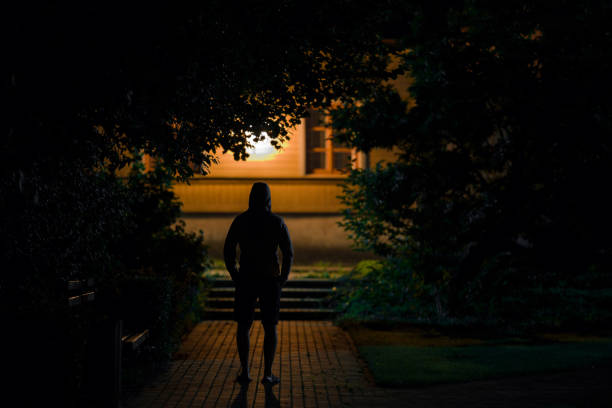 homme à capuchon restant sur le trottoir au stationnement foncé dans la nuit noire d’été. moment effrayant et atmosphère sombre. voleur regardant la maison. vue arrière. - cambriolage photos et images de collection