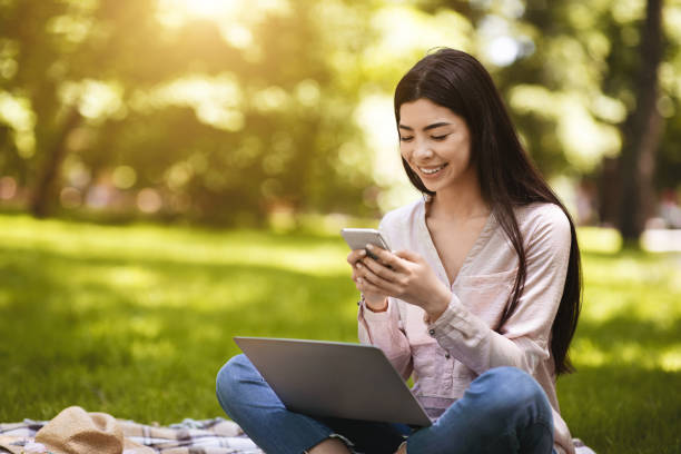 Money Transfers. Asian Girl Using Smartphone And Laptop Outdoors For Online Banking Fast Money Transfers. Smiling Asian Girl Using Smartphone And Laptop Outdoors For Online Banking, Sitting On Grass In Park, Free Space sending money stock pictures, royalty-free photos & images