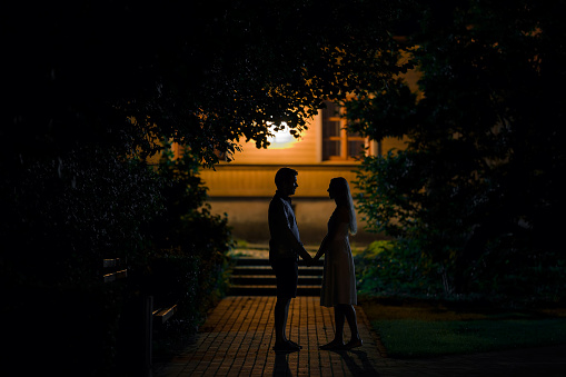 Young couple standing and staring at each other and say goodbye after dating in summer black night. Boyfriend accompanying girlfriend to home. Side view.