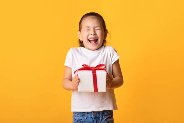 Excited asian girl holding gift box at studio Wow. Excited little kid holding gift box and screaming with joy, closed eyes and open mouth, yellow wall, copyspace christmas excitement stock pictures, royalty-free photos & images