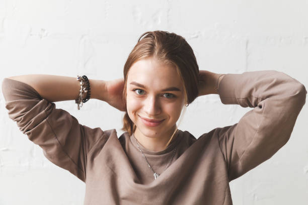 Young smiling blond girl braids her hair, close-up Young smiling blond girl braids her hair, close-up studio portrait with natural light over white wall 15 year old blonde girl stock pictures, royalty-free photos & images