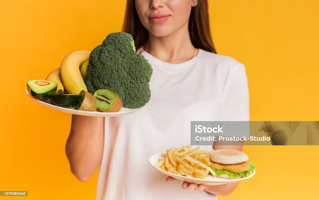 Girl choosing between organic fruits and vegetables and junk food Healthy Vs Unhealthy. Unrecognizable girl choosing between plates with organic fruits and vegetables and junk food, yellow background, cropped Dishonesty Stock Photo