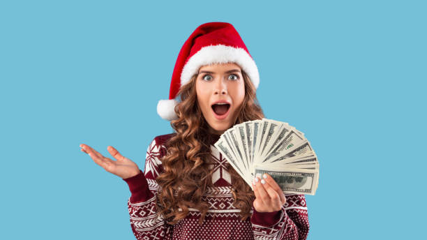 oferta especial de navidad. chica milenaria sorprendida en traje de invierno sosteniendo fan de dinero en el fondo azul - beautiful women gift christmas fotografías e imágenes de stock