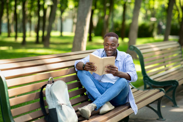 istock Positive black guy reading interesting book on bench at summer park, copy space 1270385571