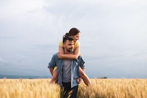Couple in a wheat field on a summer evening at sunset. Men is carrying woman on shoulders. They are having a lot of fun.