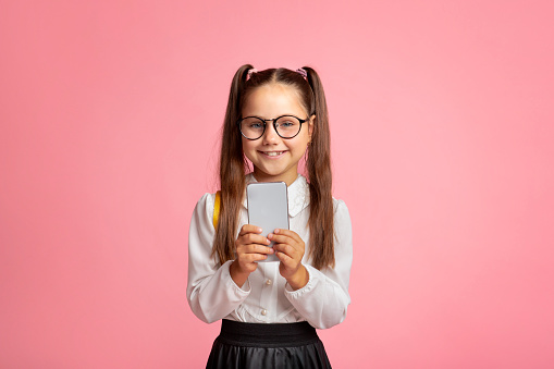 Modern technology and school. Smiling little girl with glasses holding smartphone, isolated on pink background, studio shoot, free space