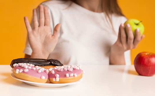 Young Woman Refusing Sweets And Choosing Healthy Fruits, Showing Stop Gesture To Plate With Donuts And Taking Apple, Cropped Image