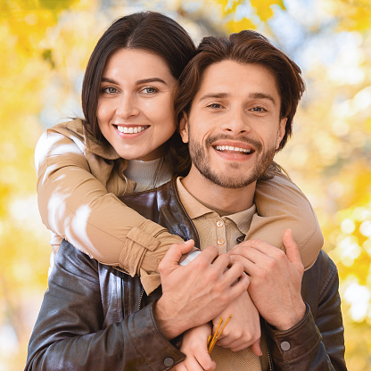 Portrait of happy young man and woman posing over golden forest, lady hugging her man from behind, close up