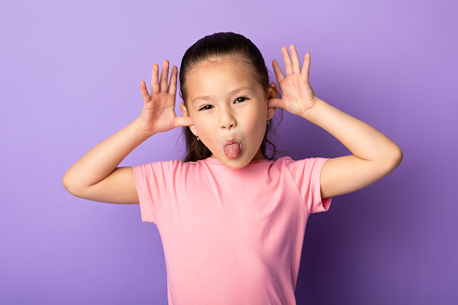 Portrait of cute asian kid sticking out her tongue and goofing at studio, isolated on pastel purple background