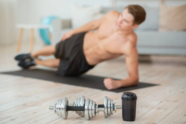 istock Dumbbells and glass for protein shake close up, guy shirtless makes side plank, on mat in living room interior 1270397078