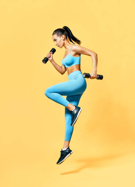sporty woman in sportswear jumping with dumbbells on yellow background - beautiful one person strength sensuality imagens e fotografias de stock