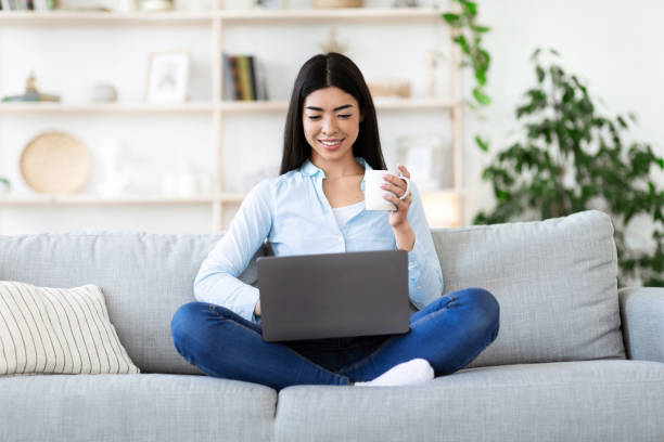 istock Leisure Concept. Asian girl relaxing on sofa, drinking coffee and using laptop 1270402300