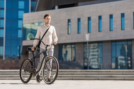 Urban ecological transport. Young smiling stylish businessman pushing bicycle while going to work near office building