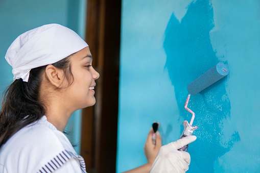 Indian Young girl painting wall with blue paint through paint roller. Home renovation.