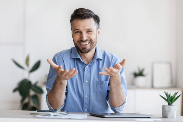 Successful handsome businessman sitting at desk in office, talking at camera Successful handsome businessman sitting at desk in office, talking at camera, sharing his experience and ideas, free space explaining stock pictures, royalty-free photos & images