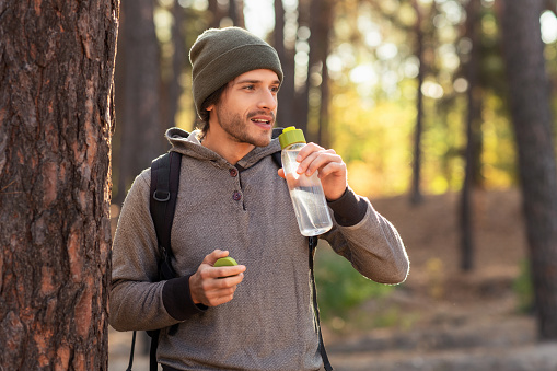 Handsome guy drinking water while walking by pine forest, copy space