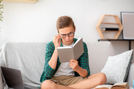 Schooler in glasses reading book at home, working on school project, empty space