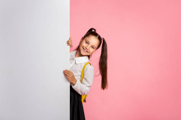 ready for lesson. cheerful schoolgirl in uniform peeps out from with white billboard - preschooler childhood outdoors cheerful imagens e fotografias de stock