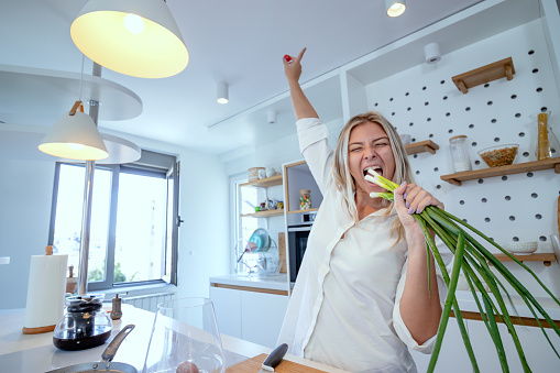 Beautiful young woman cooking in a modern kitchen. Taking a little crazy time, singing into a spring onion like into a microphone while preparing meal. Good vibes cooking concept.