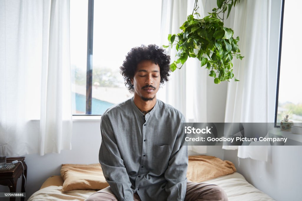 Young man sitting on his bed and meditating with his eyes closed Young African American man sitting with his legs crossed and eyes closed on his bed during a meditation session Men Stock Photo