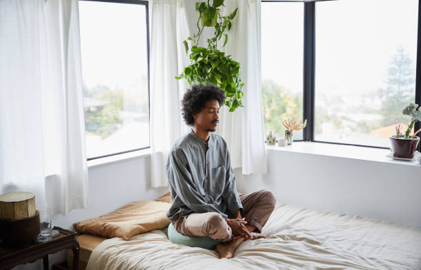 Young man sitting alone on his bed and meditating Young African American man sitting on his bed with his eyes closed and legs crossed during a meditation session meditation stock pictures, royalty-free photos & images