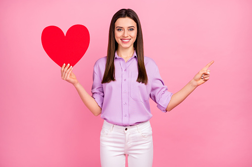 Portrait of her she nice attractive lovely pretty charming cheerful cheery, long-haired girl holding in hand big large paper heart showing advert copy space isolated on pink pastel color background
