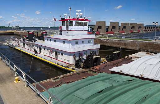 A towboat moves three rows of covered barges through a lock and past a dam on the Mississippi River.