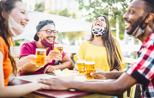 Young people drinking beer with open face masks - New normal lifestyle concept with friend having fun together talking on happy hour at brewery bar - Bright vivid filter with focus on left guy