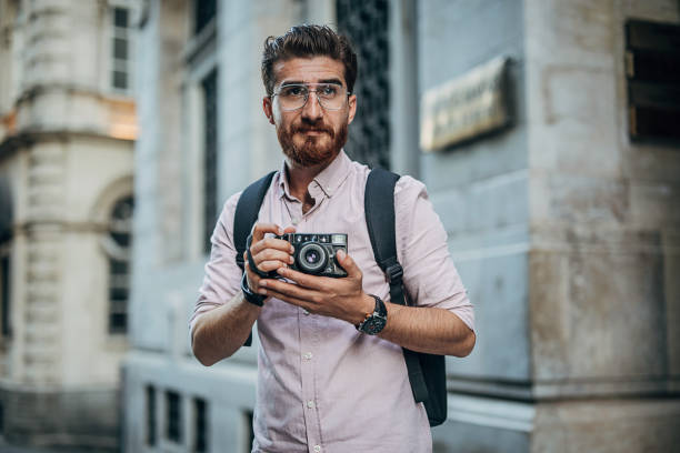 One man, modern redhead gentleman standing on the city street downtown, he is holding a camera and taking pictures.