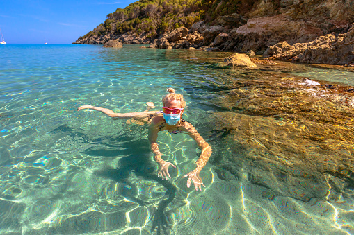 Italian girl swims in Fetovaia beach sea wearing medical mask during Covid-19. Blonde woman having a bath in Elba island sea in Coronavirus quarantine. Holidays travel in COVID pandemic Italy.