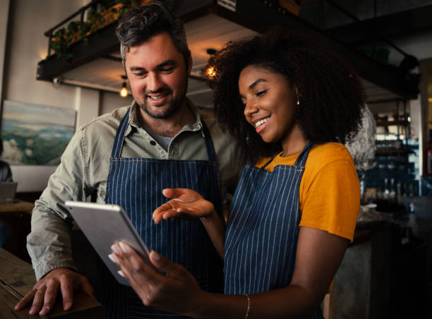 Smiling male and female colleagues discussing plans while looking at a tablet standing in coffee shop Smiling white male and ethnic female colleagues discussing business plans while scrolling on a digital tablet standing in trendy coffee shop. High quality photo african american business couple stock pictures, royalty-free photos & images
