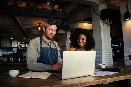 Male and ethnic female coffee shop owners smiling at laptop in coffee shop while discussing financial plans . High quality photo