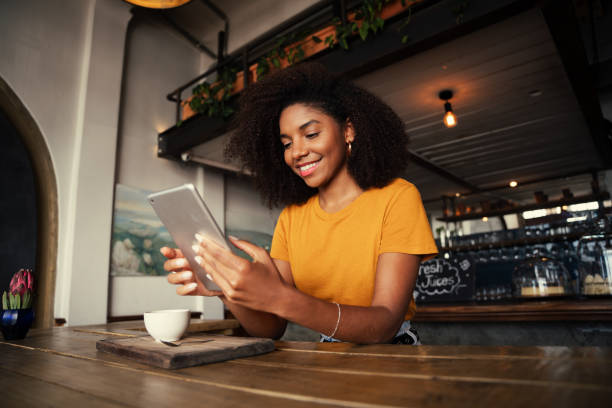 Low angle shot of beautiful African American female holding digital tablet sitting at funky tearoom with hot latte Low angle shot of unique African American woman searching on digital tablet sitting at funky tearoom with hot latte. High quality photo cafe tablet stock pictures, royalty-free photos & images