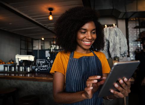 Excited African American waitress wearing apron scrolling on digital tablet in coffee shop. High quality photo