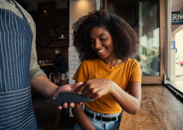 Smiling mixed race woman with afro happily paying for coffee at coffee shop Smiling mixed race female with afro happily tapping card machine after paying for coffee at coffee shop. High quality photo tapping stock pictures, royalty-free photos & images