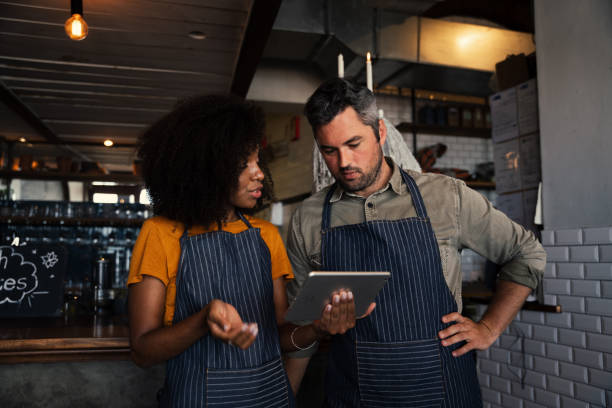 istock Curious colleagues wearing aprons, discussing finances while looking at tablet standing in coffee shop 1270459723