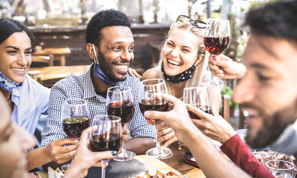 friends toasting red wine at outdoor restaurant bar with open face mask - new normal lifestyle concept with happy people having fun together on warm filter - focus on afroamerican guy - wine dinner party drinking toast imagens e fotografias de stock