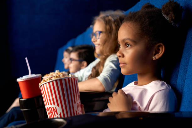 African girl with friends in cinema. Beautiful concentrated african girl with funny hairstyle watching movie in cinema. Adorable little female kid sitting with friends, eating popcorn and smiling. Concept of entertainment, leisure. cute entertainment stock pictures, royalty-free photos & images