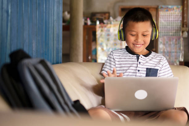 apprendimento remoto online, istruzione a distanza e concetti di homeschooling. ragazzo di scuola asiatico preteen ragazzo in cuffia utilizzando computer portatile sul divano nella casa rurale rustica durante la pandemia di covid-19. - asian country foto e immagini stock