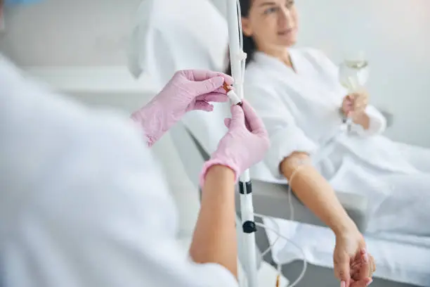 Dark-haired spa client with a glass of drink in her hand looking away during the intravenous therapy