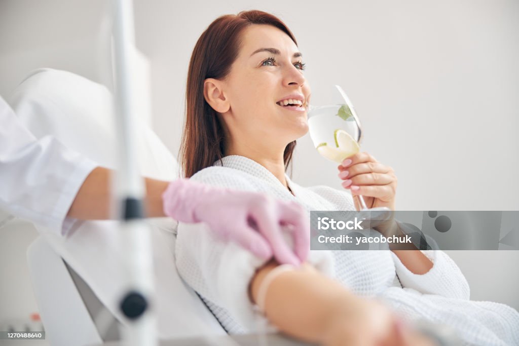 Attractive dark-haired woman smiling during the intravenous therapy Joyous female patient drinking a healthy beverage during a medical procedure in a beauty clinic IV Drip Stock Photo