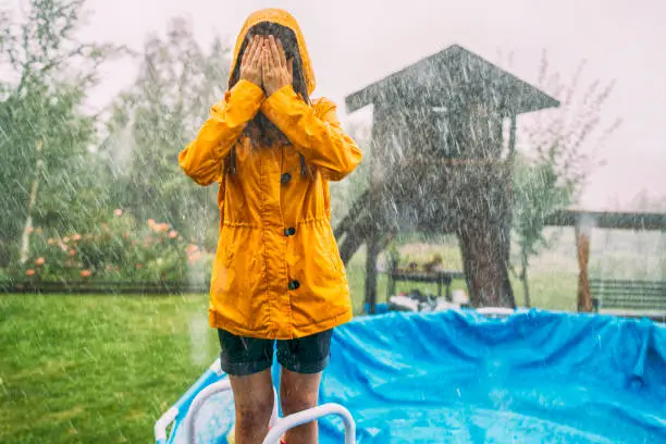 Young girl standing on the pool stairs under a heavy rain with hands covered the face