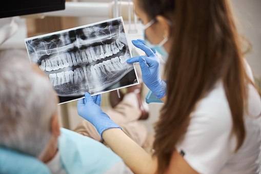 Qualified young female dentist holding an x-ray image of teeth and showing it to her elderly patient