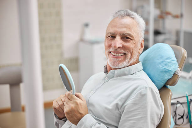 Happy eldelry man sitting in a dental chiar Cheerful patient sitting in a dental chair and holding a mirror while smiling at the camera dentists chair stock pictures, royalty-free photos & images