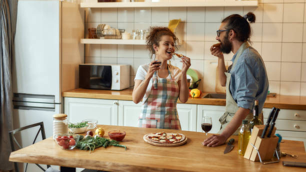 divertirsi. giovane uomo e donna in grembiule che mangiano pizza appena sforata e bevono vino mentre si trovavano in cucina. amore, concetto di relazioni - italian culture women looking at camera cheerful foto e immagini stock