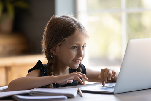 Cute small girl child sit at desk look at laptop screen play online game on gadget at home, smart little kid browse internet study distant on modern computer device, children and technology concept