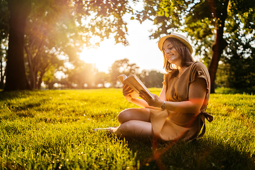 Young Caucasian smiling beautiful woman reading book on meadow at sunset.